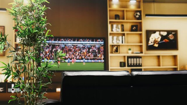 Soccer game broadcast on TV in empty apartment living room before fans arrive with cozy sofa, plant and television set screen creating comfortable home viewing setup for sports event.