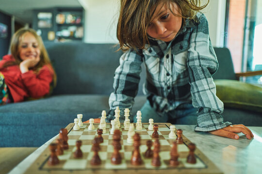 Boy studying a chess board while his brother watches from the sofa