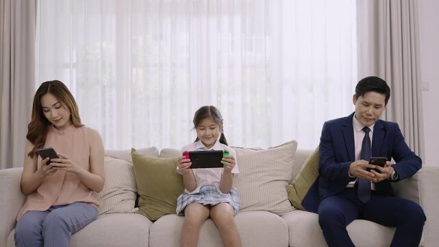 Asian family devices addicted to and overuse social media, so they are not paying attention to each other father and mother playing smartphones while daughter play game console on sofa in living room.