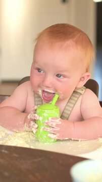 Vertical screen. Baby with red hair engages with a green sippy cup, making a mess with food on a high chair in a well-lit indoor environment