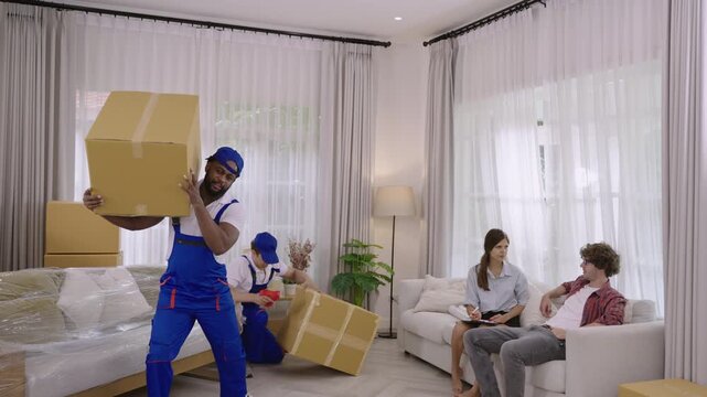 Two professional movers in uniform are moving furniture and cardboard boxes during a home relocation, while a young couple of homeowners, holding a clipboard, are checking for delivery at their home. 