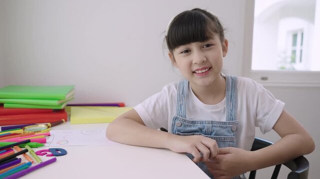 Cute Asian girl student sitting at desk is looking at the webcam while talking to computer camera and video calling with a remote teacher for online learning at home. Homeschooling education concept. 