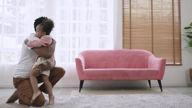 A cute African American child daughter is running and smiling to embrace her father while sitting and waiting on the carpet floor, happy and bonding in the living room at home. Father's Day concept. 
