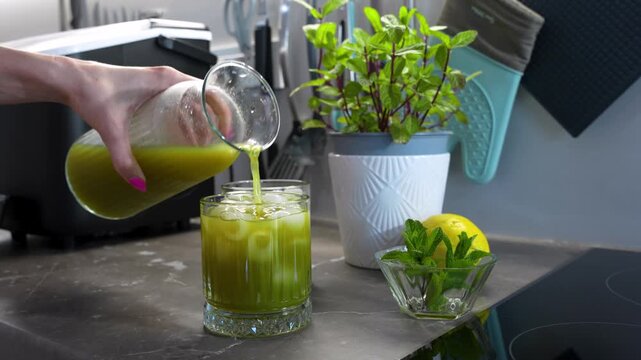 Homemade mint lemonade being poured over ice in a glass, with fresh lemon and mint on a kitchen counter for a refreshing summer drink theme, close-up view