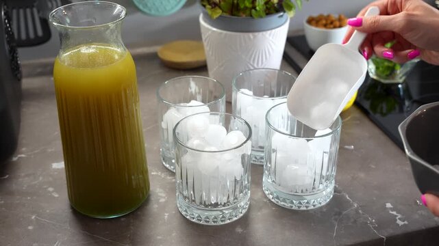 Close up of a hand using a white scoop to fill crystal glasses with ice cubes next to a pitcher of Arabian mint lemonade. Authentic home kitchen setting for summer wellness and refreshing drinks.