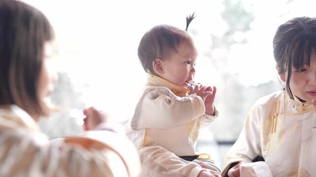 Soft light bathes a tender family moment. Baby reaches with curious hands, eyes wide with wonder. Two women smile, watching the child with affectionate joy. Scene suggests quiet domestic warmth, calm