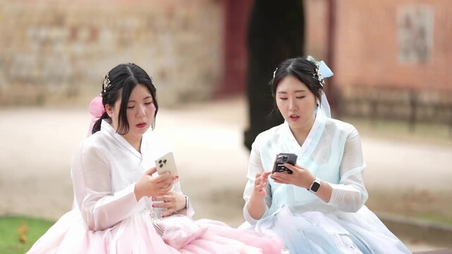 Two women in traditional Korean dresses sit on grass. They use smartphones while seated near an old stone building. Their focused expressions suggest quiet engagement or leisurely browsing