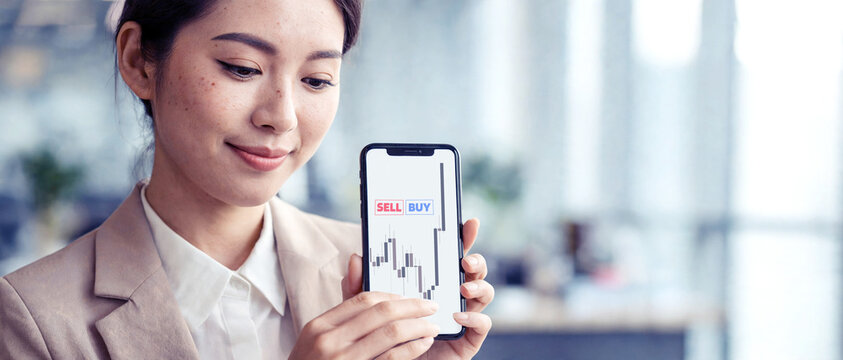 Mujer de negocios asi&aacute;tica muestra gr&aacute;fico de velas de Forex y botones de comprar y vender en su tel&eacute;fono m&oacute;vil. Profesional sonriente haciendo trading en una oficina moderna.