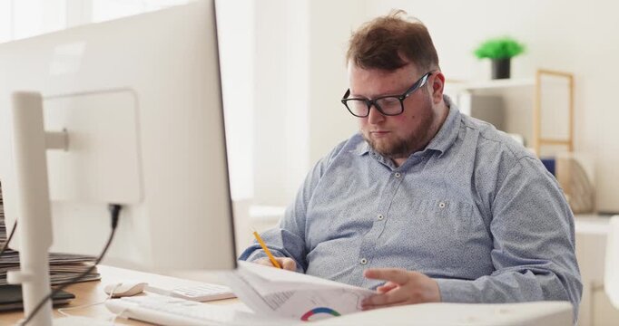 Man accountant in office doing budget analysis. He reviews finance paperwork at the computer, checking numbers and managing expenses while stressed. Concept shows stress and responsibility.