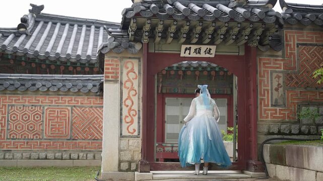 Woman in a flowing blue gown stands before a traditional Korean gate. She gazes upward, perhaps in quiet contemplation. The gate features intricate tilework and carved wood