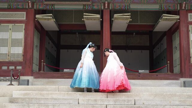 Two women in colorful traditional dresses stand on stone steps. Their heads are bowed, showing quiet reverence. They face a grand, red-painted temple entrance. The scene evokes cultural heritage