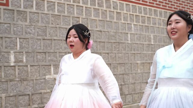 Two women in hanbok dance joyfully. One wears pink, the other light blue. They smile, move with energy. Their traditional attire contrasts the stone wall. Perfect for cultural, festival