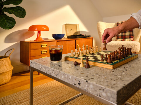 A man's hand moves a wooden piece on a chessboard next to a glass of iced coffee on a terrazzo table in a cozy mid-century modern living room.