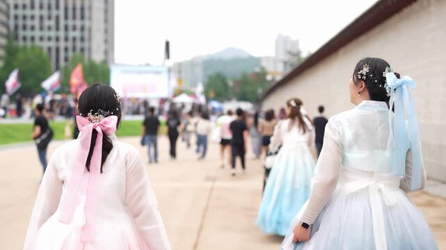 Two girls in traditional Korean dresses walk away from camera. Pink and light blue ribbons frame their backs as they stroll. Other people move around them, blurred in background