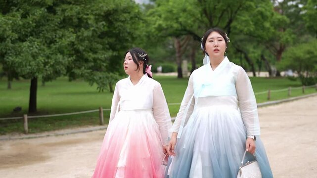 Two women in colorful hanboks walk side by side. Their smiles radiate joy as they stroll through a park. Lush green trees frame the path behind them. The soft breeze lifts their flowing skirts gently