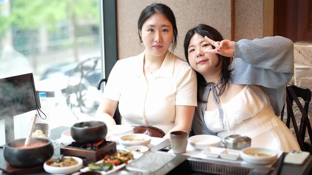 Two women sit at a table, smiling. They make peace signs with their fingers. The table holds multiple bowls and a stone pot. They eat together in a restaurant. Natural light streams through the window