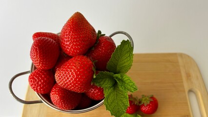 Fresh red strawberries with mint leaf in a small metal colander on a wooden cutting board © LD