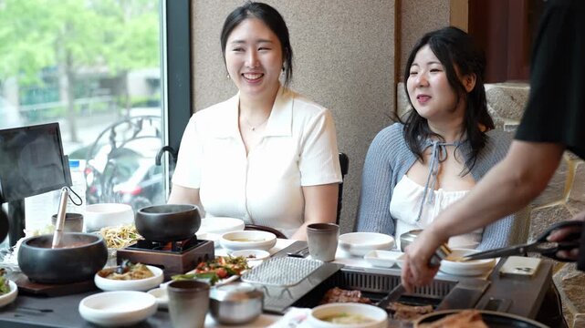Two women laugh during a lively meal. Server adds food to their shared table. The scene suggests a cozy, shared dining experience. They enjoy the warmth and company. The table holds various dishes