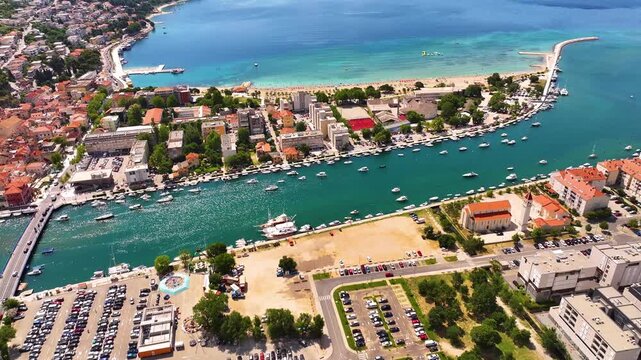 Omis shows a waterfront area with boats in the water. Nearby buildings and a beach are visible. A parking lot is located on the right side. People are present along the shore.