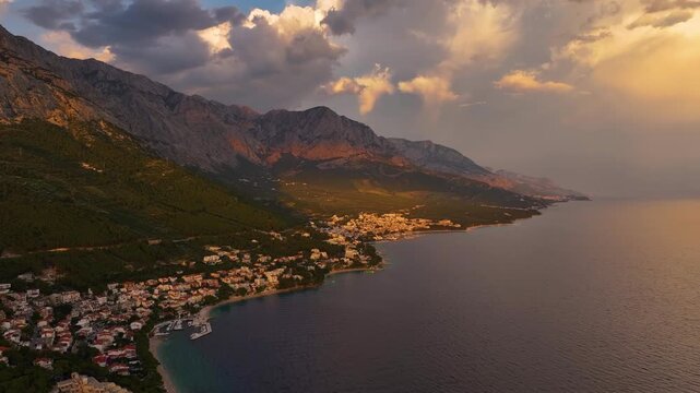 Aerial view of Brela Croatia shows the town by the sea. Mountains are visible in the background. Sunlight reflects on the water. Clouds fill the sky.