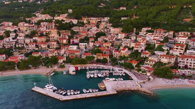 Brela features a coastal area with various residential buildings. Boats are docked in the marina. The beach area is visible. Green hills are in the background.