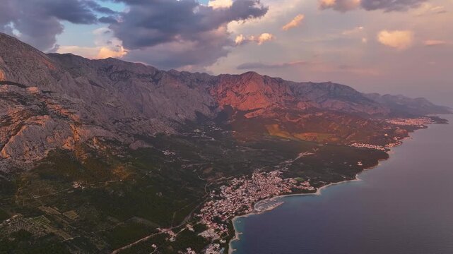 Brela shows an aerial view of the coastline and mountains. The village is near the water. Mountains are visible in the background. The sun is setting.