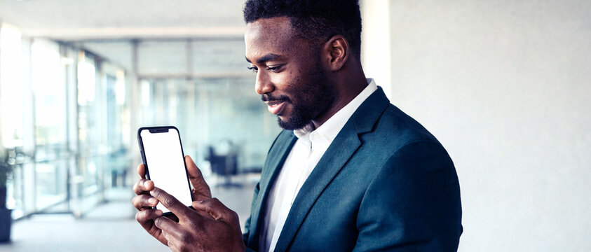 Hombre de negocios afroamericano sonr&iacute;e interactuando con la pantalla blanca vac&iacute;a de su tel&eacute;fono m&oacute;vil en la oficina. Maqueta publicitaria para aplicaciones, marketing y servicios digitales.