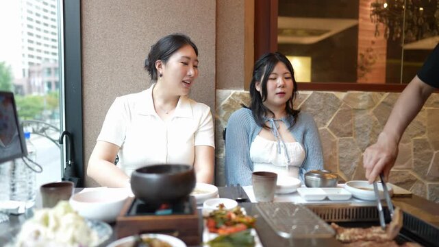 Two women enjoy Korean-style hot pot dining together. They laugh, smile, and chat while sharing food. Hand serves meat from a sizzling grill. The setting is cozy, with stone walls and soft lighting