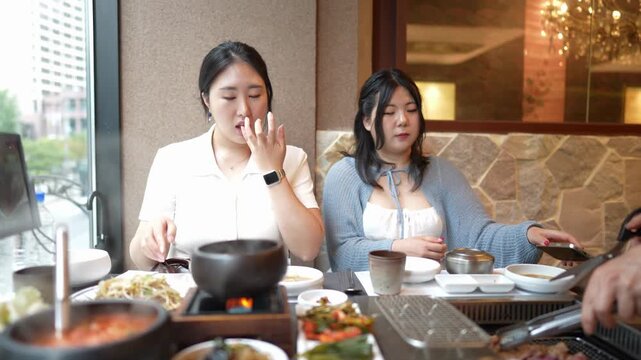 Two women enjoy Korean barbecue together. One looks at her phone while the other eats. They sit at a table with hot pots and grilled meat. The restaurant has warm lighting and stone walls