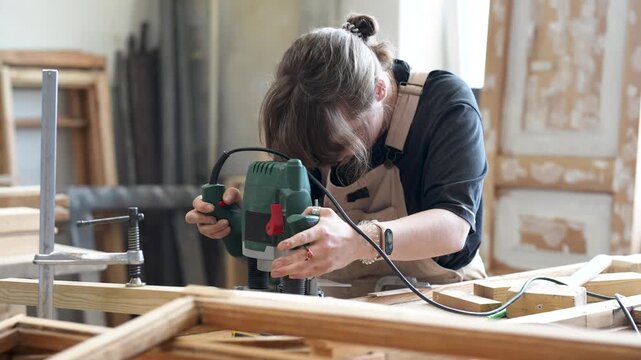 Concentrated woman restorer working with handheld plunge router, cutting off layer from board surface, female master engaged in working process, precisely levels wooden material with instrument.