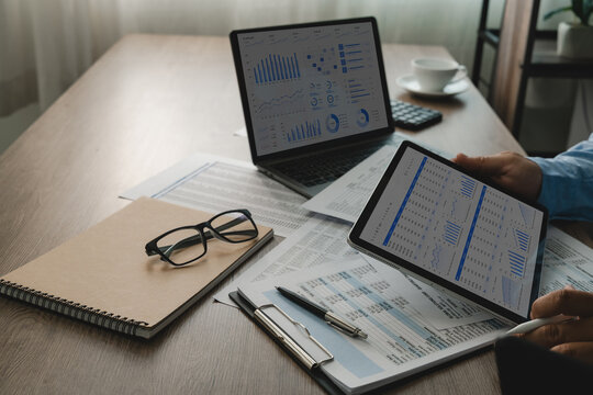 Laptop and tablet display financial dashboards above printed reports on a desk, while hands review charts and tables for financial data analysis, reporting, and business performance tracking.