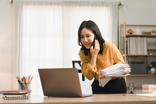 Asian woman holding documents while talking on phone and working at a laptop in office