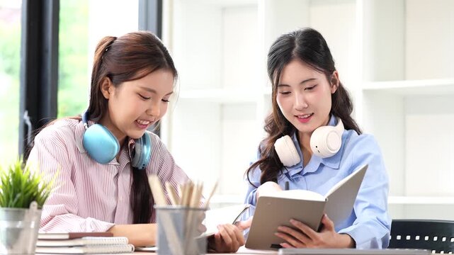 Two happy young Asian female students with headphones studying together, smiling and writing in a notebook at a desk. Education and lifestyle concept.