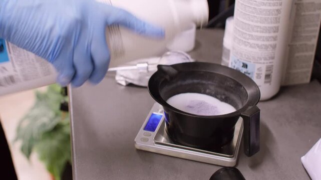 Measuring hair dye. Hairstylist mixing color. Professional woman hands in gloves pouring white developer using bottle in beauty salon.