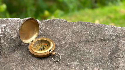 Naklejka premium A brass compass with its lid open rests on a stone surface against a backdrop of blurred greenery. Conceptually, it suggests travel, navigation, exploration, adventure, and finding one's bearings.