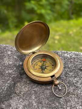A classic metal compass with its lid open, resting on a stone against a backdrop of blurred greenery. A symbol of travel, freedom, exploration, new horizons, independence, and inspiration.