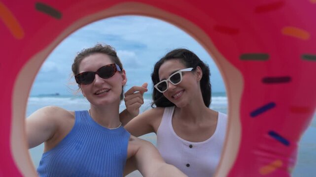 Beach summer vacation selfie woman friend smile sunglasses float ocean joyful candid at sandy shore, finger heart pose under blue sky and soft wave, pink donut ring vibe, playful carefree laugh
