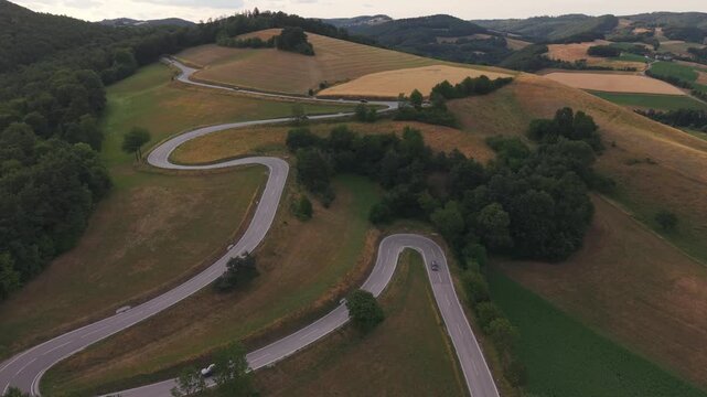A breathtaking drone view of a winding mountain road with multiple hairpin bends crossing rolling hills, forests, and patchwork farmland.