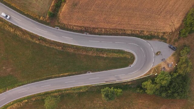 A top-down drone view of a sharp hairpin bend on a winding rural road with cars driving through an agricultural landscape.