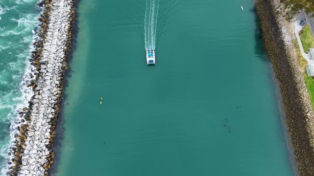 The drone leads a boat from high altitude through the harbor toward the Pacific Ocean. A graceful shot captures the beauty of the calm greenish blue waters below as the boat slowly glides along the sr