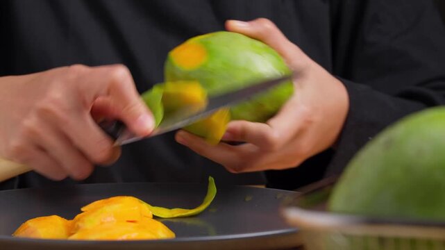 Hands peel mango with sharp knife over dark plate