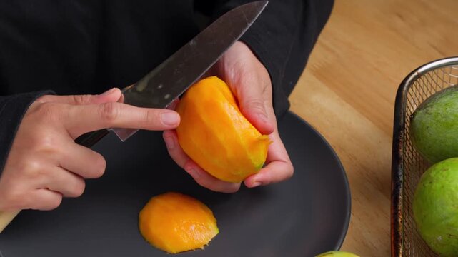 Hands peel mango with sharp knife over dark plate