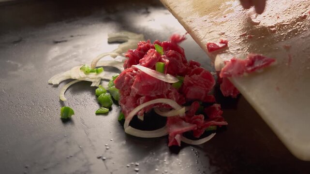 Hand adds chopped meat to hot griddle with onions and green pepper. Cooking scene captures fresh, raw ingredients being prepared