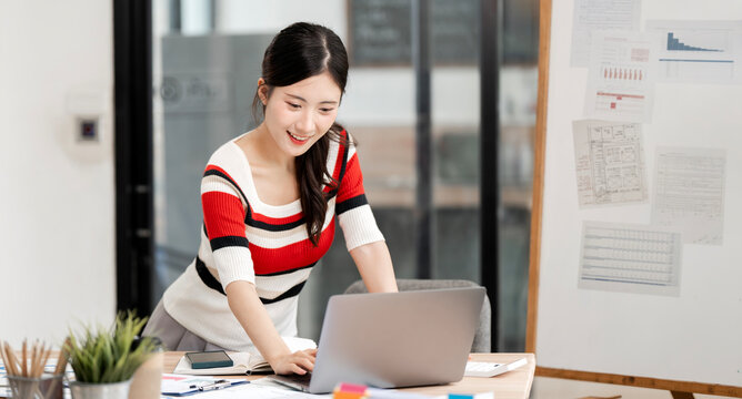 Happy Young Asian Woman Leaning Over Desk and Working on Laptop