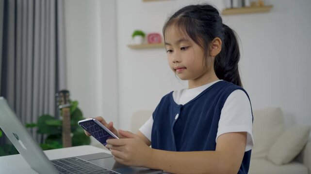Young girl using smartphone while sitting at table with laptop in living room focused on screen flexible modern technology usage at home study and communication