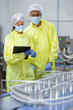 factory worker in yellow protective suit and medical mask holding clipboard near metal conveyor production line during manufacturing inspection process inside modern clean room facility