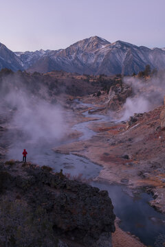 Solitary hiker stands experiencing profound wonder watching dense geothermal steam rise near majestic snow mountain peak in sierra nevada valley landscape enjoying peaceful dawn purple sky