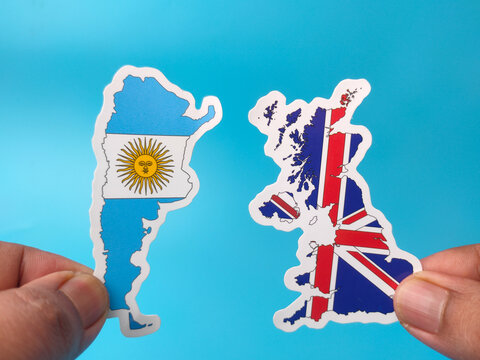 Two hands hold up country map stickers of Argentina and the United Kingdom decorated with their national flags