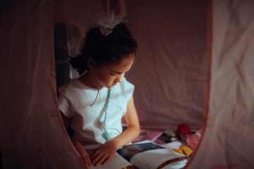  Little Girl Reading Book Inside Cozy Tent In Dim Light. Curious child sits in dark play teepee enjoying storybook during cozy evening indoors  © nicoletaionescu