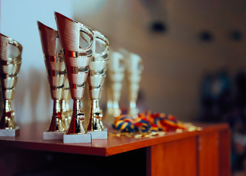Row Of Shiny Silver Trophies Close Up Before Awards Ceremony. Metallic prize cups standing on wooden table with blurred medals in background